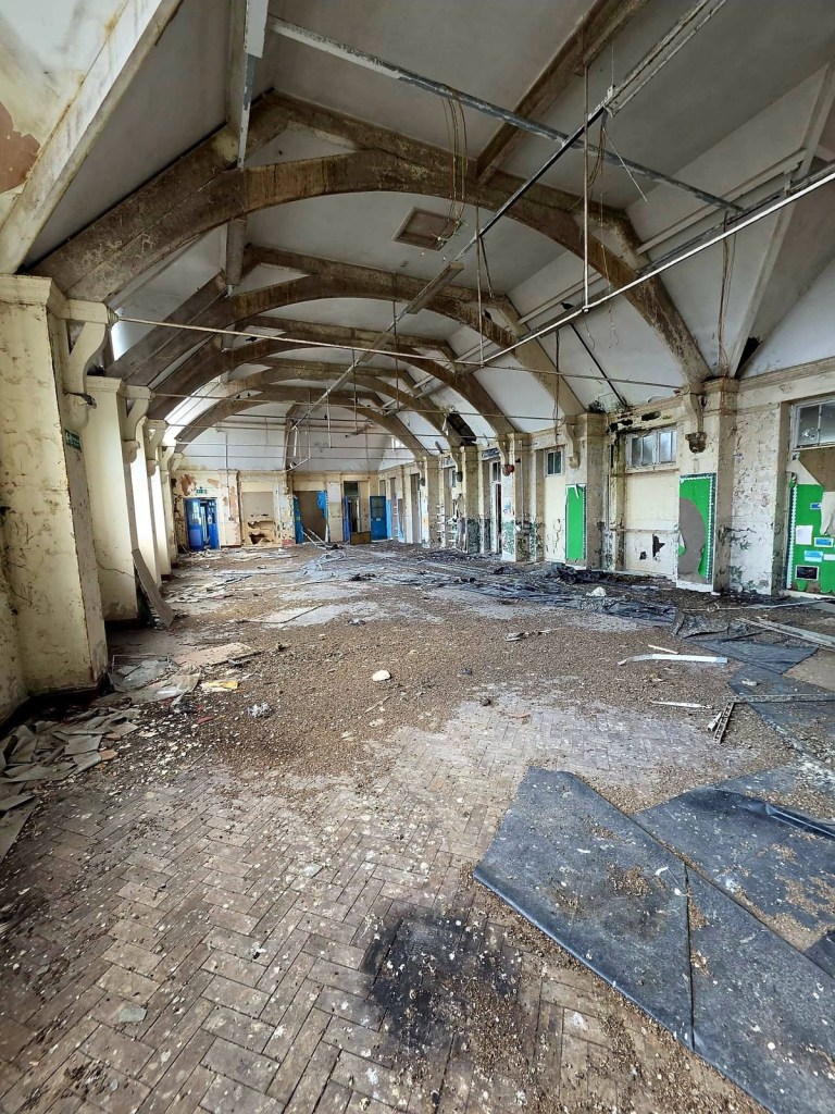 Large empty school hall with arched concrete roof beams, debris across the floor and blue classroom doors along the walls
