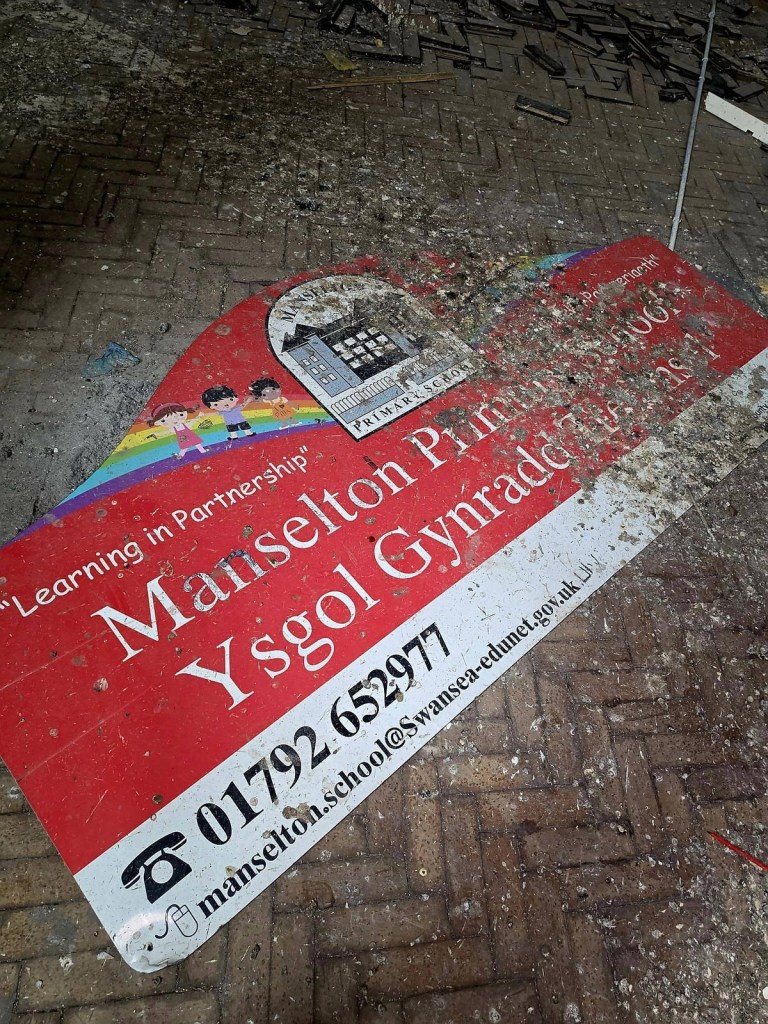 A fallen Manselton Primary School sign lying on a debris-covered parquet floor inside the abandoned school