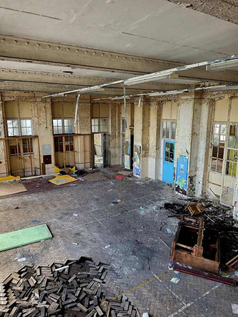 View from above of a derelict school hall with a smashed upright piano and piles of broken parquet blocks