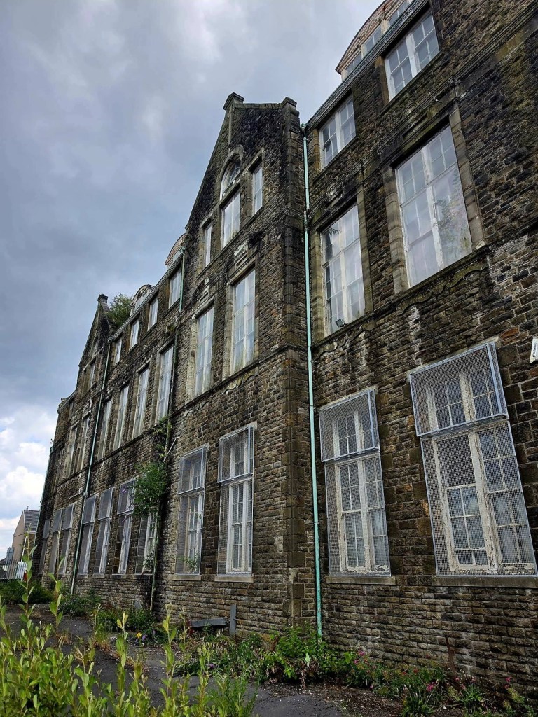 Side elevation of the Victorian stone school with caged lower windows and plants growing from the masonry