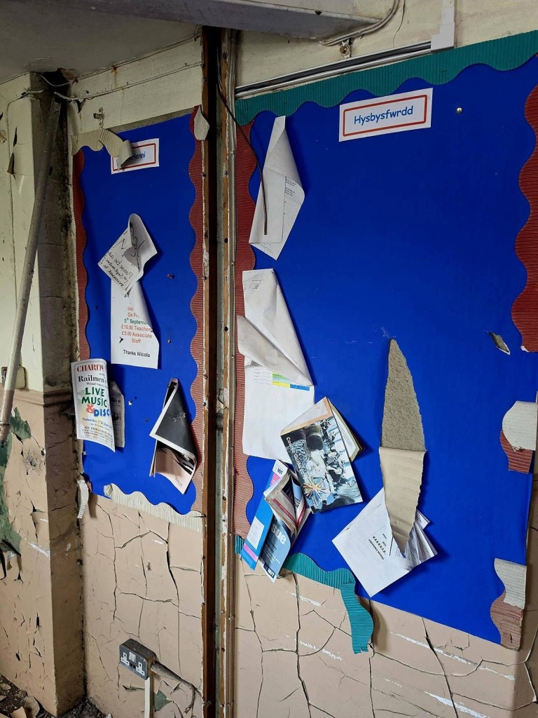 Staff noticeboard marked Hysbysfwrdd with curling notices and leaflets still pinned in a derelict school corridor