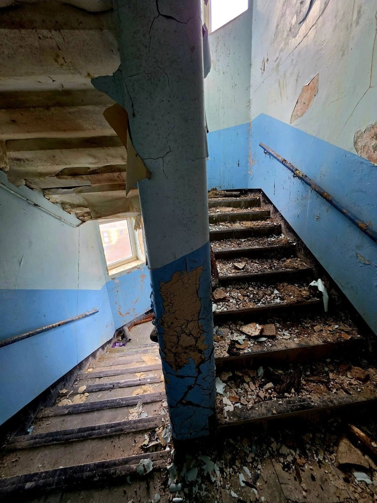 Derelict school staircase covered in fallen plaster with peeling blue and white paint