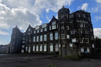 Exterior of the abandoned former Manselton School in Swansea, a three-storey Victorian stone building with turrets, boarded ground-floor windows and weeds growing through the yard