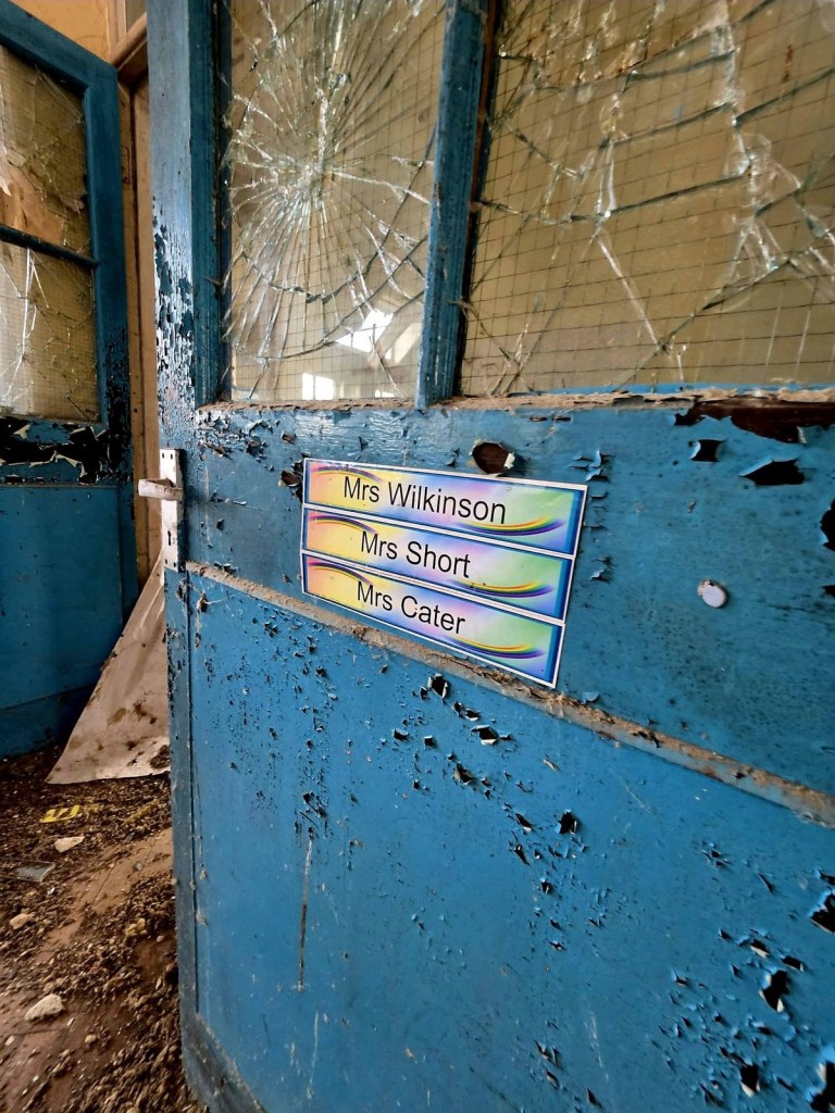 Peeling blue classroom door with smashed wired glass and rainbow name plates for three teachers