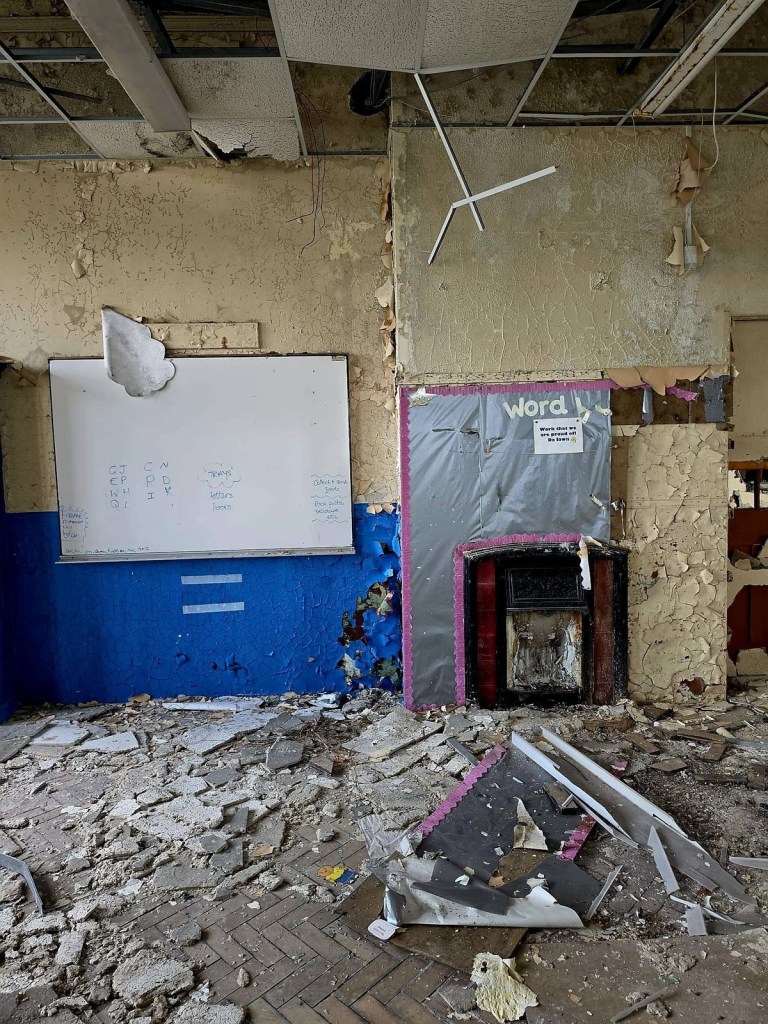 Classroom whiteboard with handwritten packing-up notes next to a Victorian fireplace and a Word Wall display in a derelict classroom