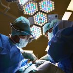 Two surgeons in blue scrubs, gowns and masks operate under bright theatre lights