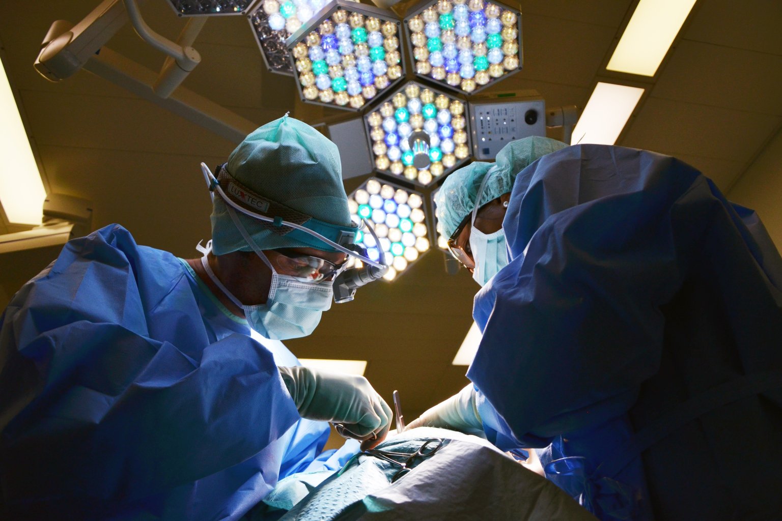 Two surgeons in blue scrubs, gowns and masks operate under bright theatre lights