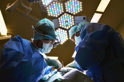 Two surgeons in blue scrubs, gowns and masks operate under bright theatre lights