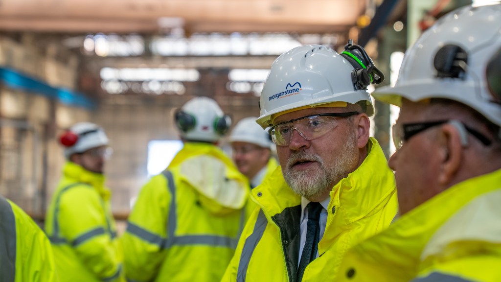 A man with a grey beard in safety glasses and a white Morganstone hard hat inside the steelworks, with other workers in hi-vis behind him