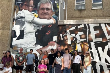 A large mural on a building wall on Salubrious Place, Swansea, showing a grandad and his young granddaughter in football stands with the words Feels Like Home, with a crowd gathered in front