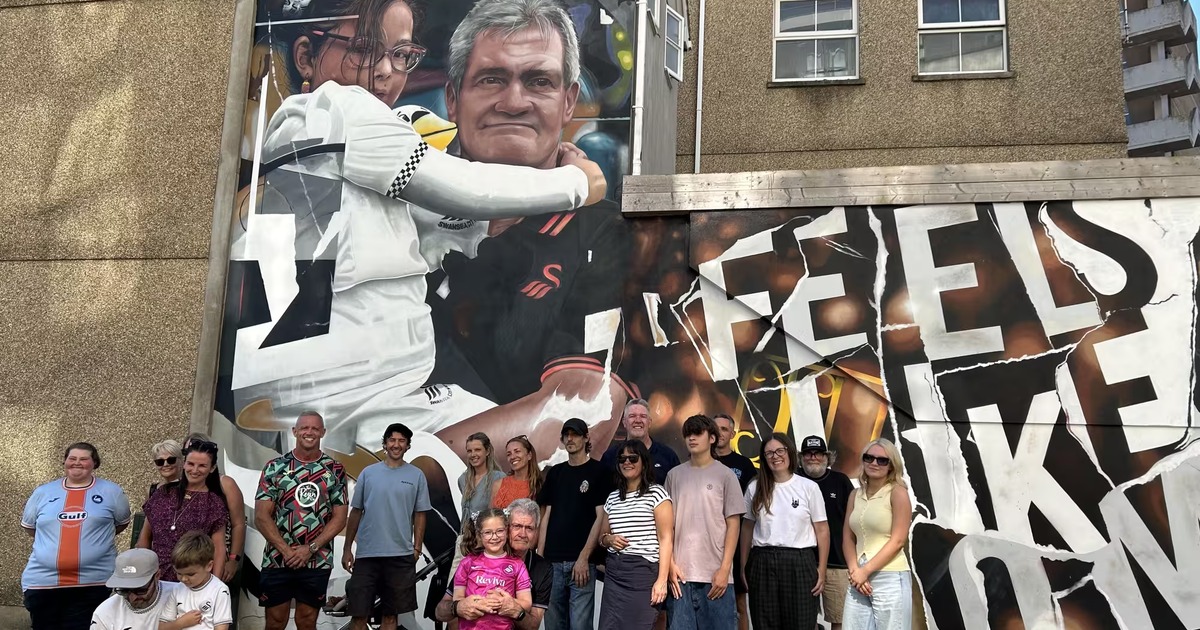 A large mural on a building wall on Salubrious Place, Swansea, showing a grandad and his young granddaughter in football stands with the words Feels Like Home, with a crowd gathered in front