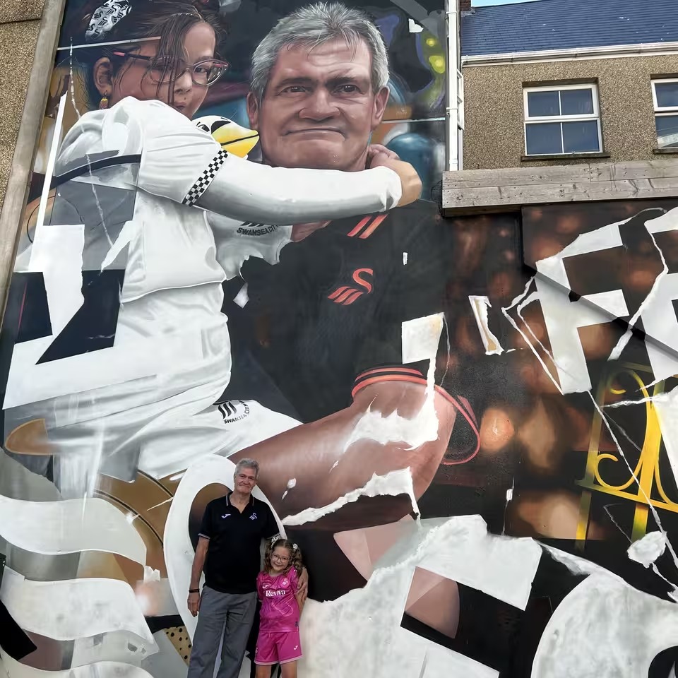 Close-up of the mural showing an older man and a young girl side by side in the stands, with the real Salvatore Rullo and Rosa Allen standing in front of it