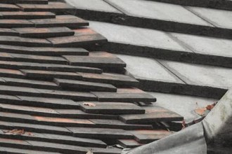 A roof with rows of dark tiles sitting unevenly and out of line, with visible gaps between courses.