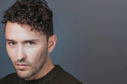 Richard Mylan, a man with dark curly hair and stubble in a dark green knitted jumper, looking directly at the camera against a grey background.