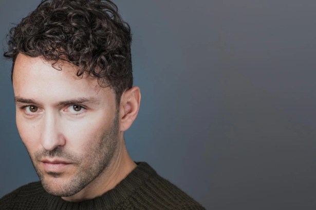 Richard Mylan, a man with dark curly hair and stubble in a dark green knitted jumper, looking directly at the camera against a grey background.