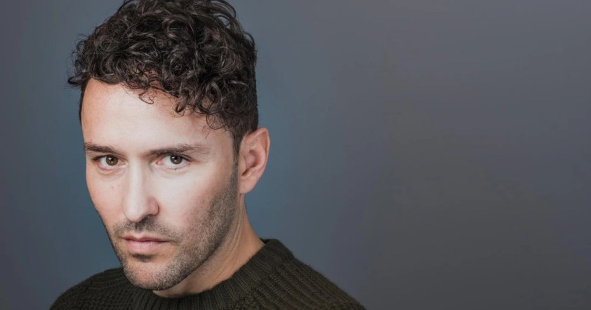 Richard Mylan, a man with dark curly hair and stubble in a dark green knitted jumper, looking directly at the camera against a grey background.