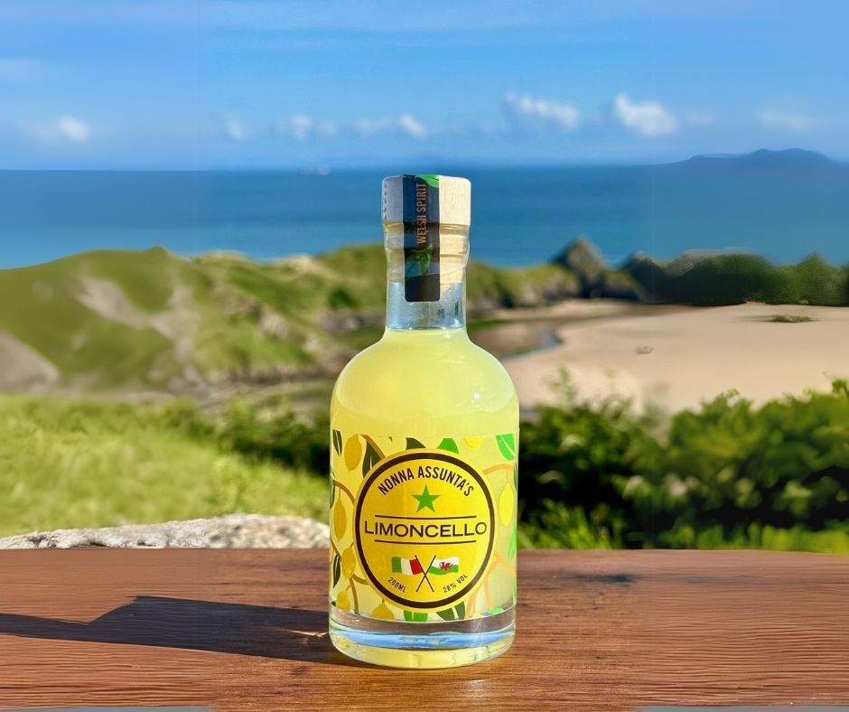A bottle of Nonna Assunta's Limoncello on a wooden table with a sandy Gower bay and headland behind