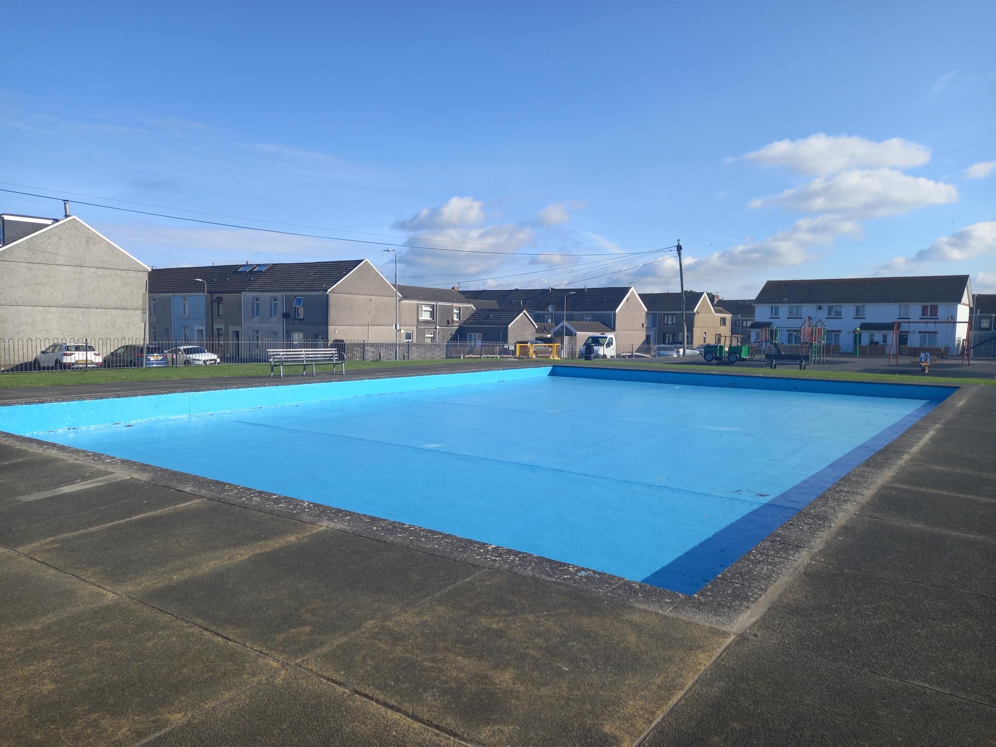 The empty, freshly painted blue paddling pool at Crown Park, Llanelli, with houses and a playground behind