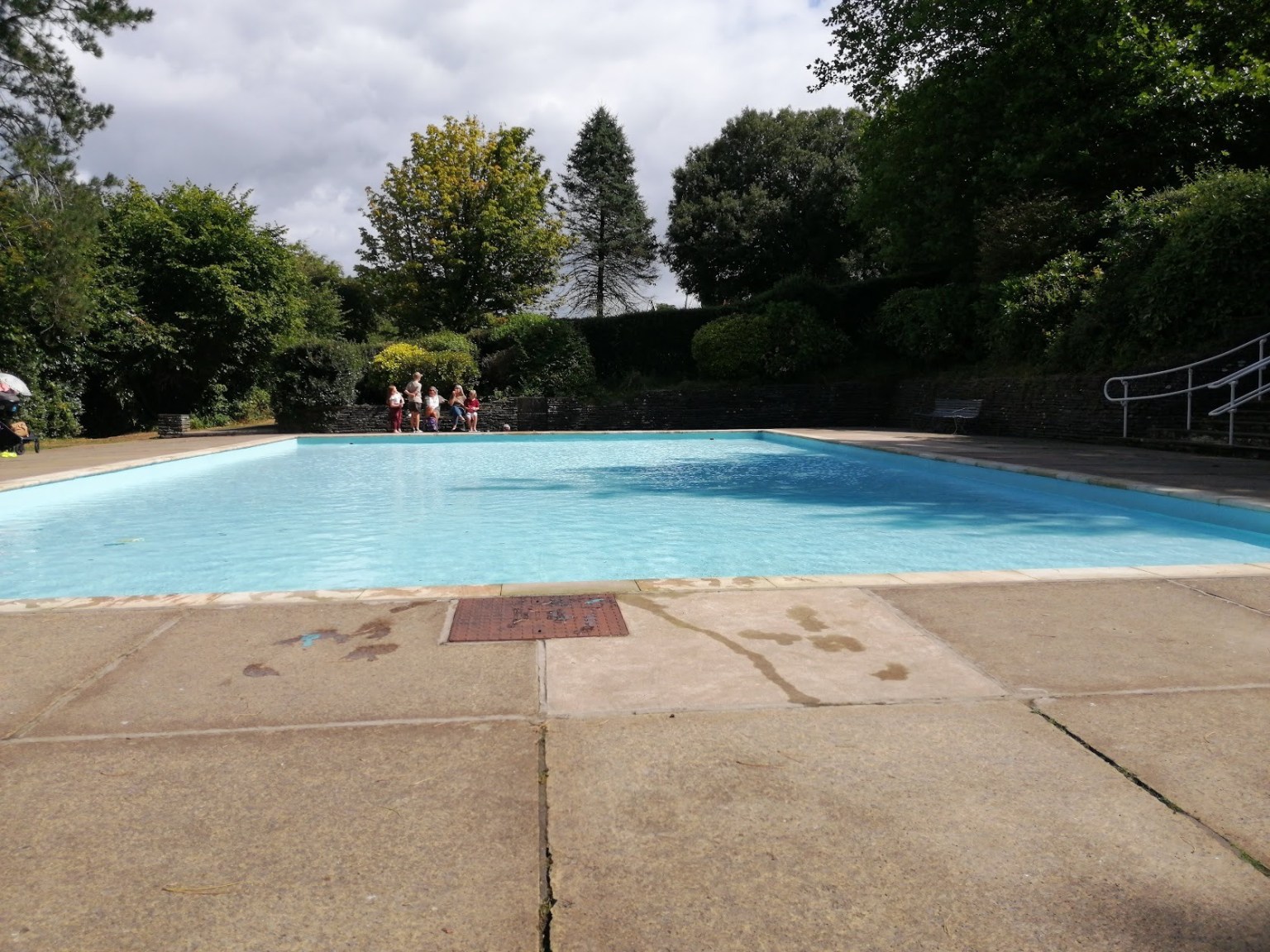 The filled paddling pool at Parc Howard, Llanelli, surrounded by trees with families sitting at the poolside