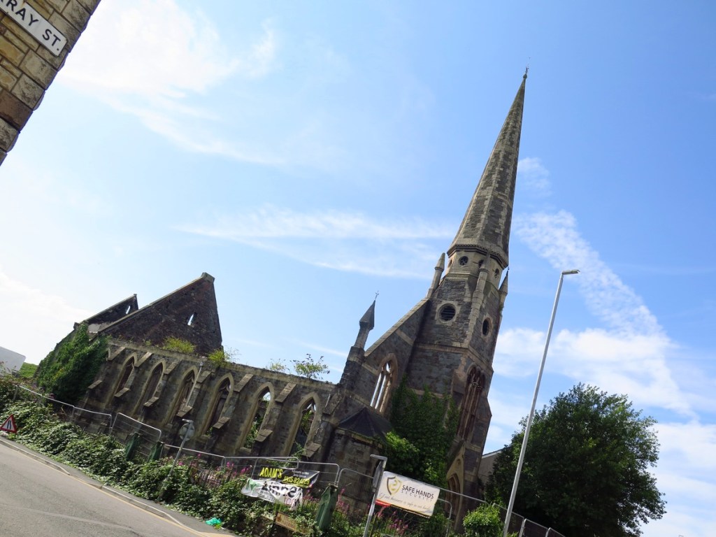 A ruined Gothic chapel with a tall spire seen from street level, its windows empty and walls overgrown, behind metal security fencing.