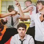 Five actors in school uniform in a changing room scene, one sitting calmly while the others gesture around him. Optional: full poster inline at the end, same credit.
