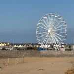 A large white observation wheel towering over Sandy Bay beach in Porthcawl, with houses and the seafront wall behind