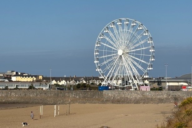 A large white observation wheel towering over Sandy Bay beach in Porthcawl, with houses and the seafront wall behind