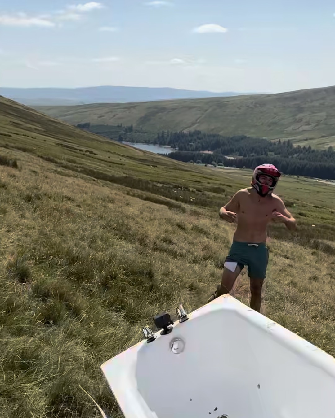 A man in a red motocross helmet stands on a hillside beside an upended white bathtub, a reservoir and forestry in the valley behind.