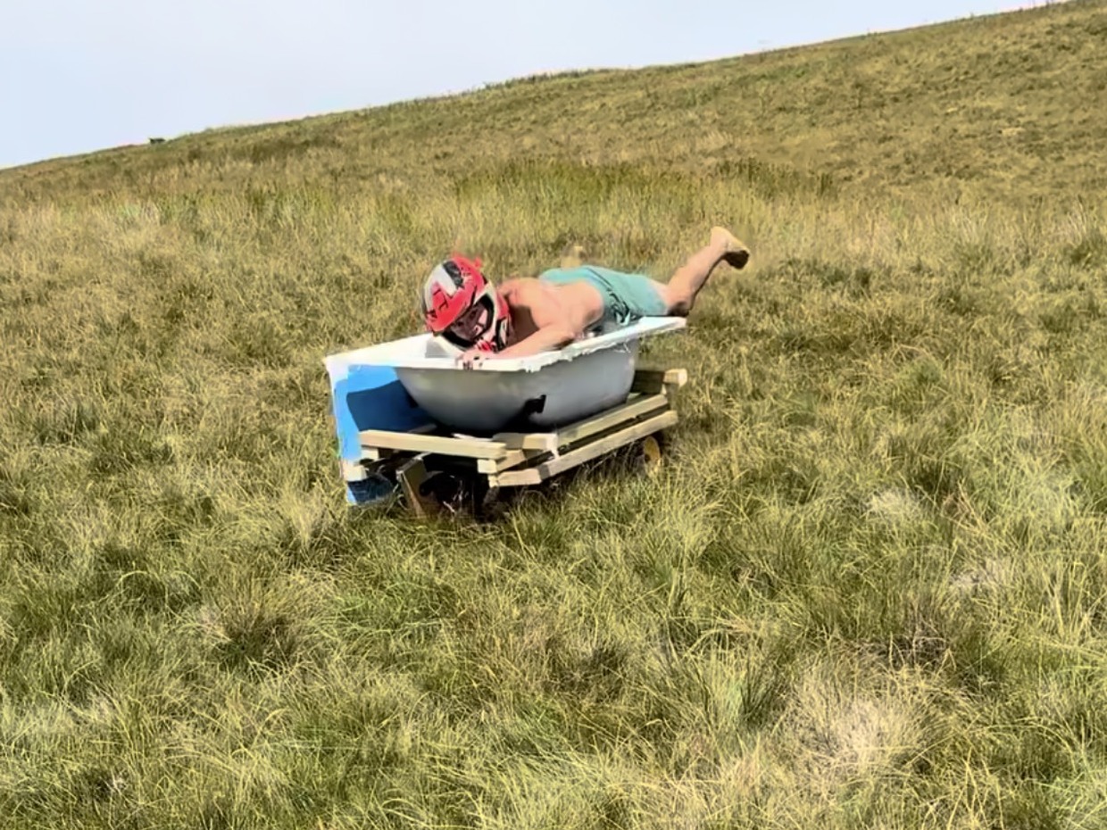 A man in a red helmet lies in a bathtub mounted on a wheeled wooden frame, part-way down a wide grassy slope.