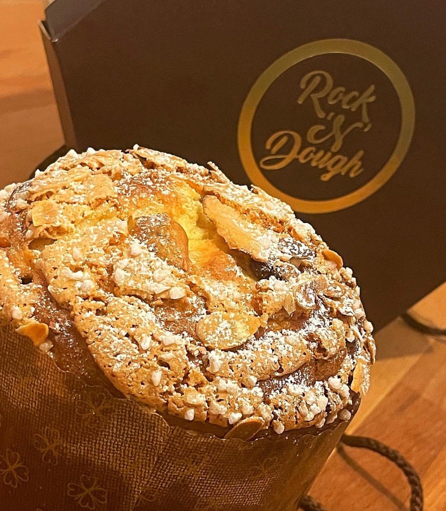 A sugar-dusted almond-topped panettone in its baking wrapper, with a Rock'n'Dough branded box behind