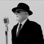 Roger Taylor in a black hat, sunglasses and suit standing at a vintage microphone against a plain background