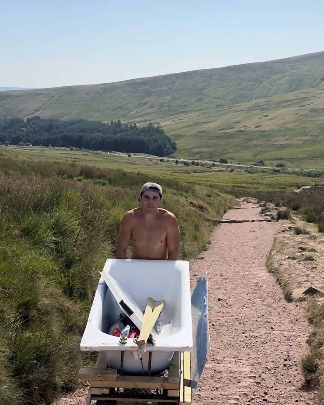 A shirtless man pushes a broken white bath on a wheeled wooden frame down a stony mountain path, valley and main road below.