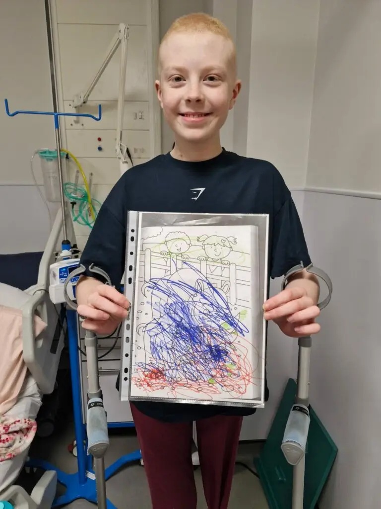 Soffia smiling and holding up a colouring picture in a hospital room