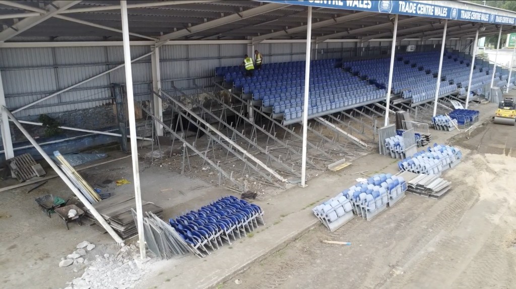 Workers in high-vis removing blue seats from the part-stripped main stand at St Helen's, with removed seats stacked in piles below
