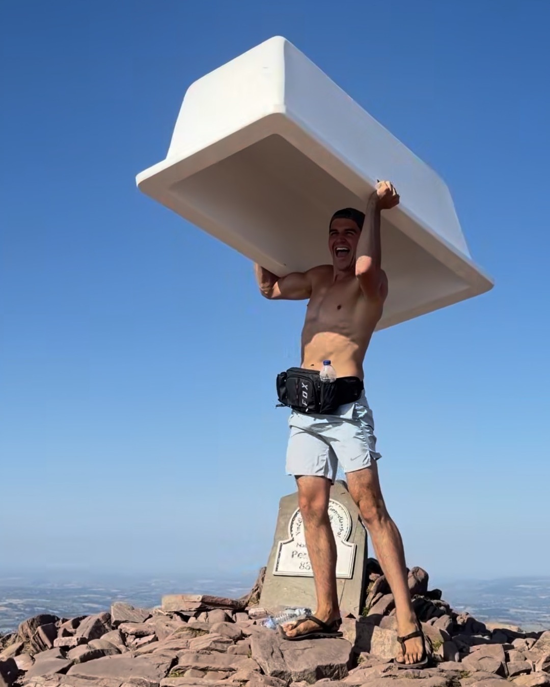A shirtless man in shorts and flip flops holds a white bath above his head on the stone summit platform of Pen y Fan beside the summit marker.