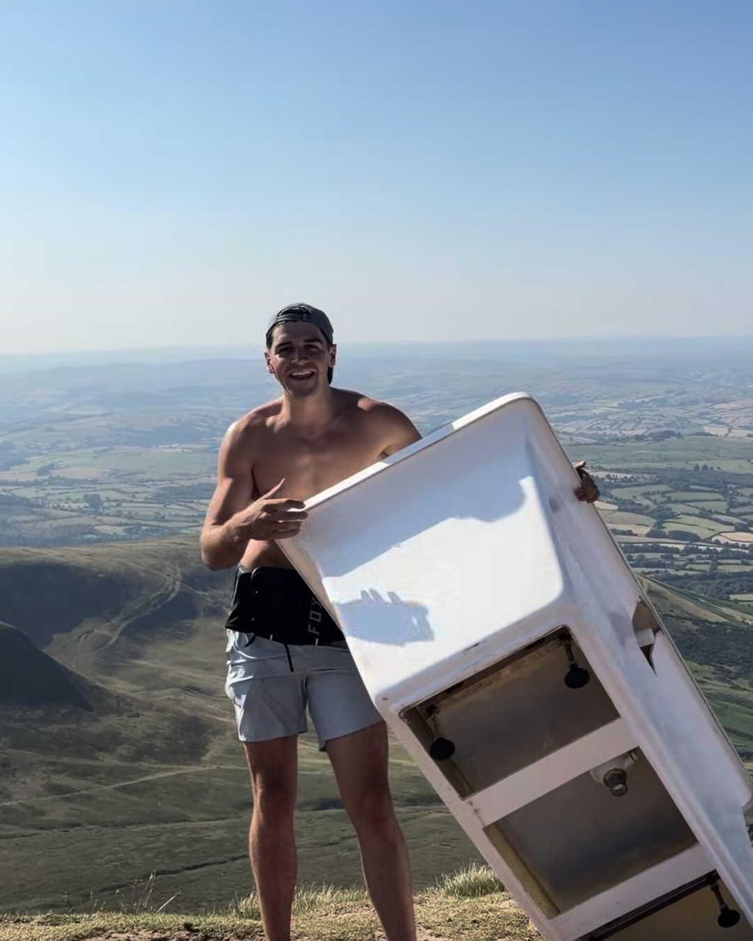 A shirtless man stands on the summit edge holding a white bath tipped on its side, patchwork farmland far below.