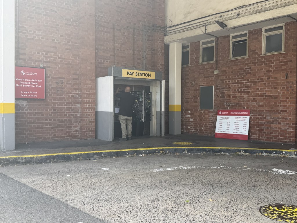 A man working inside the open pay station kiosk at Orchard Street car park, with a red Swansea Council tariff board propped nearby