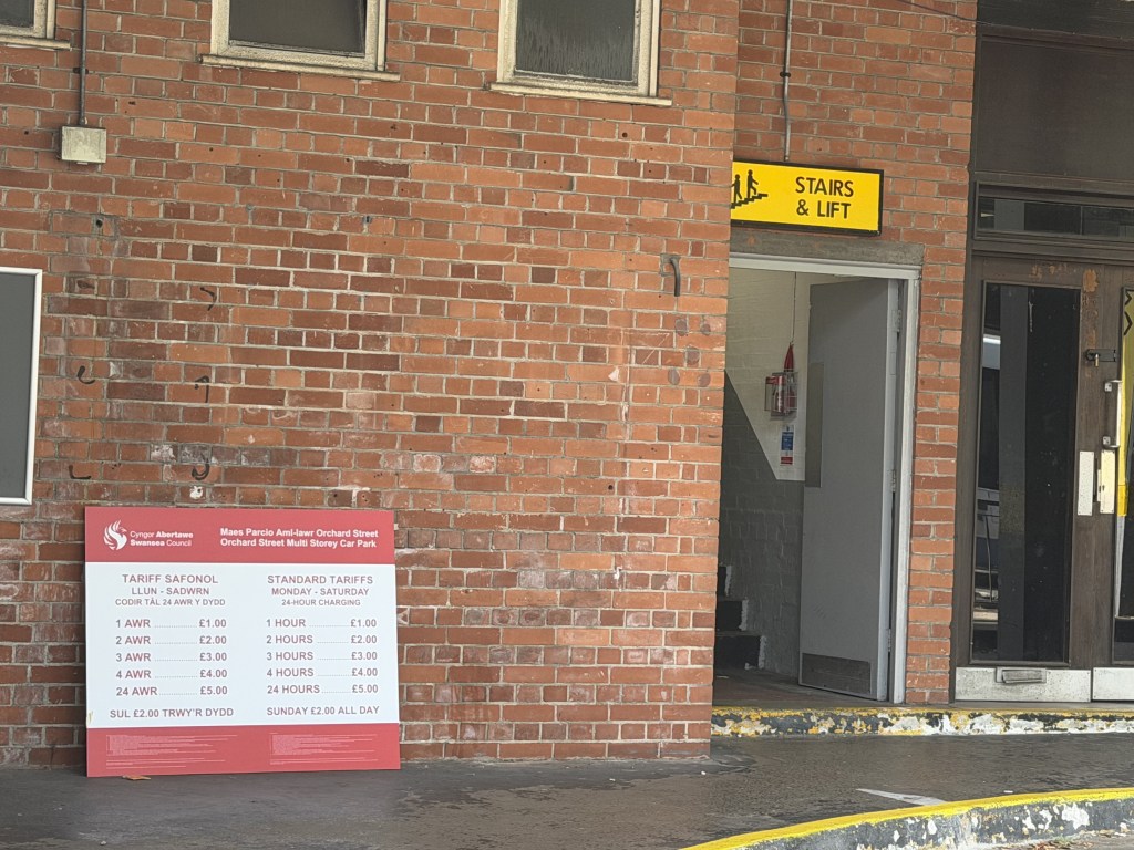 A red Swansea Council tariff board propped against a brick wall at Orchard Street car park listing charges of £1 for one hour and £5 for 24 hours