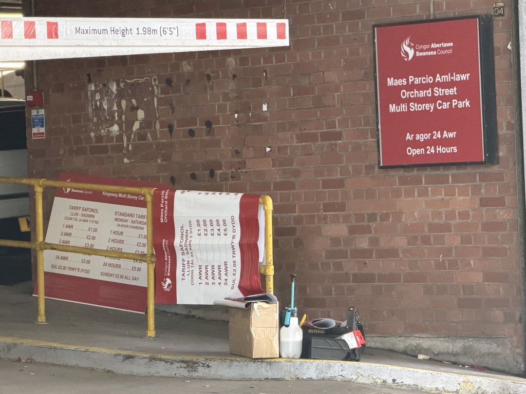 Inside the Orchard Street car park entrance, a new red Swansea Council sign on the brick wall beside the faded outline where the NCP sign was removed, with tariff boards propped against a barrier