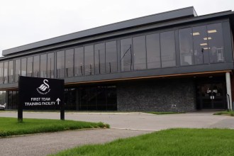 The exterior of Swansea City's first team training facility at Fairwood, a modern glass-fronted building with the club crest on a sign outside.