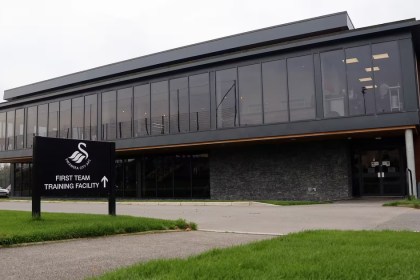 The exterior of Swansea City's first team training facility at Fairwood, a modern glass-fronted building with the club crest on a sign outside.