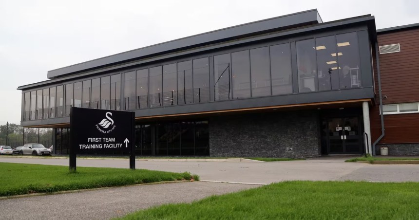 The exterior of Swansea City's first team training facility at Fairwood, a modern glass-fronted building with the club crest on a sign outside.