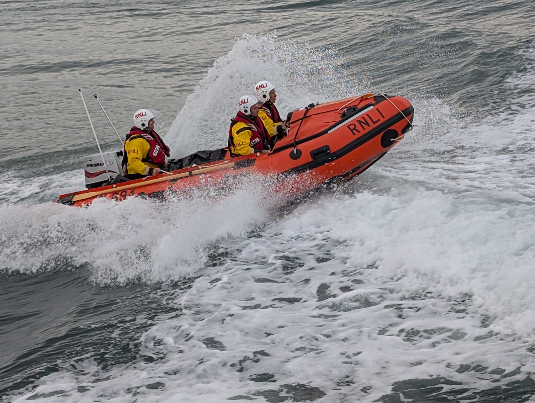 Three RNLI crew in an orange D-class inshore lifeboat cutting through waves
