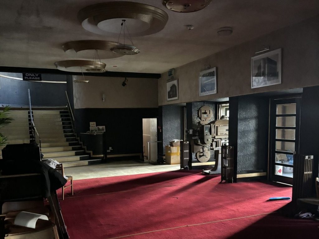 The dim foyer of a closed cinema with red carpet, a staircase, circular art deco ceiling features and a vintage film projector.