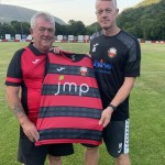 Two men hold up a red-and-black hooped Trefelin BGC shirt on the pitch at Ynys Park