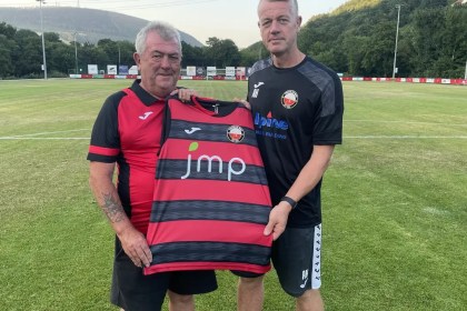 Two men hold up a red-and-black hooped Trefelin BGC shirt on the pitch at Ynys Park