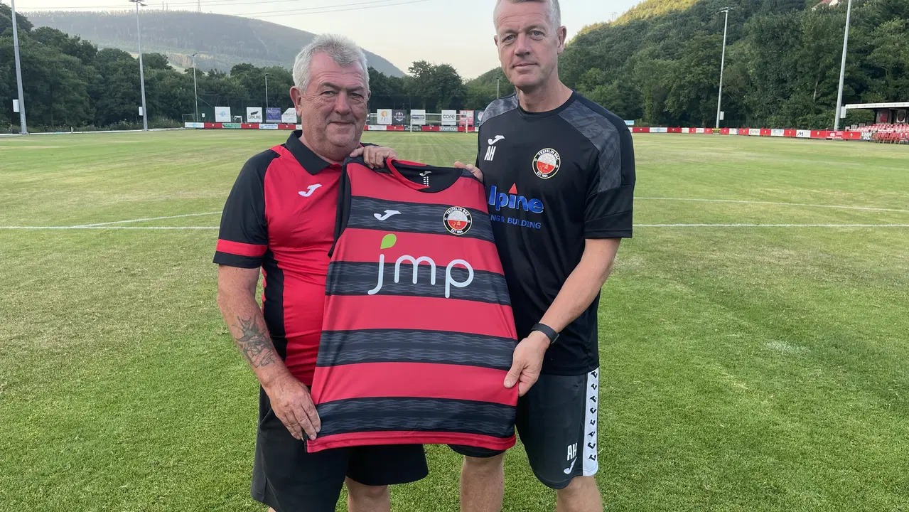 Two men hold up a red-and-black hooped Trefelin BGC shirt on the pitch at Ynys Park