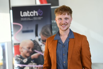Owain Wyn Evans, smiling in an orange blazer and striped shirt, in front of a Latch charity banner.
