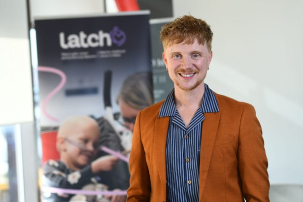 Owain Wyn Evans, smiling in an orange blazer and striped shirt, in front of a Latch charity banner.