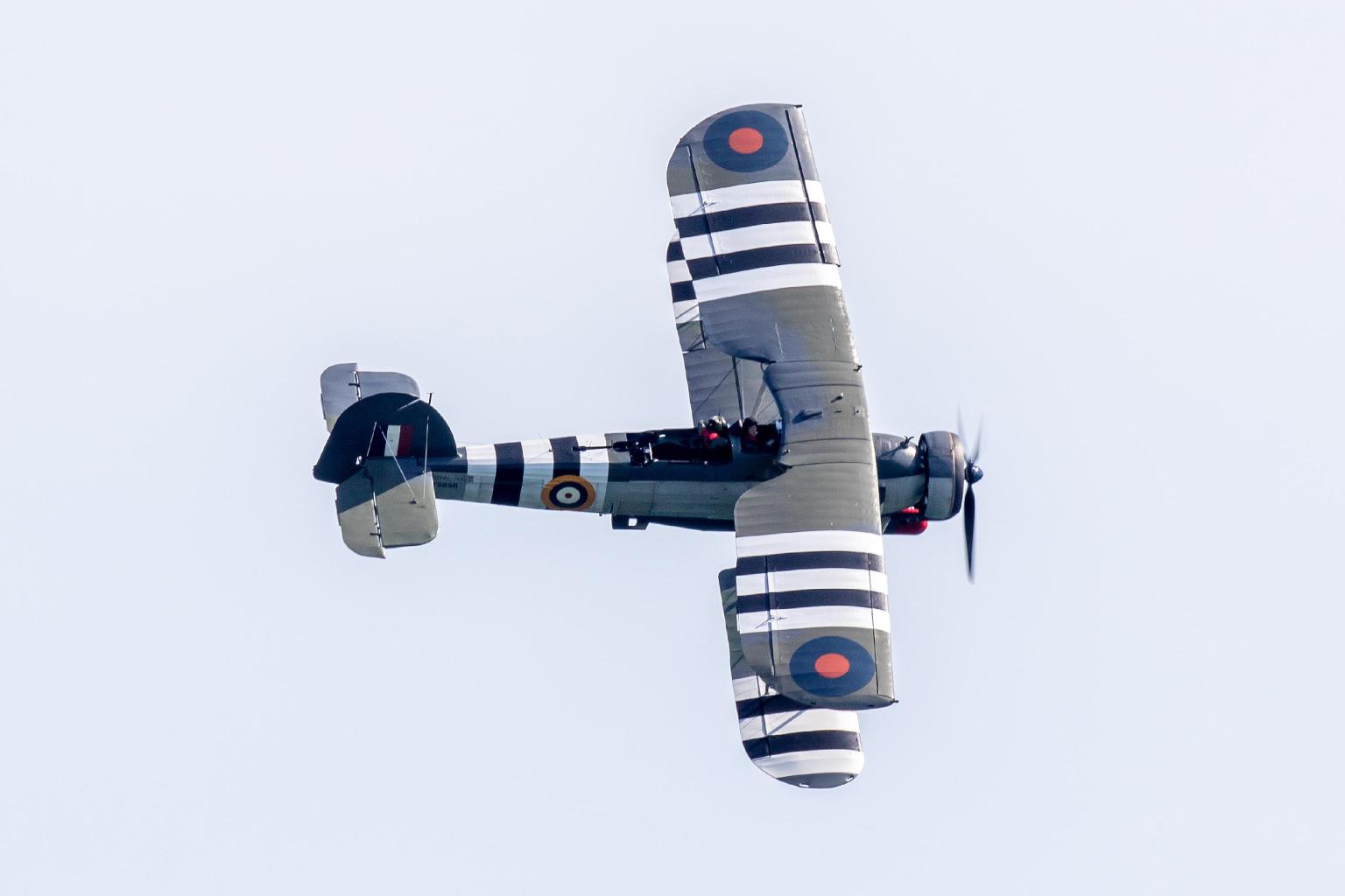 The Fairey Swordfish biplane in Royal Navy colours with black and white D-Day invasion stripes, banking against a pale sky with two crew visible in the open cockpit